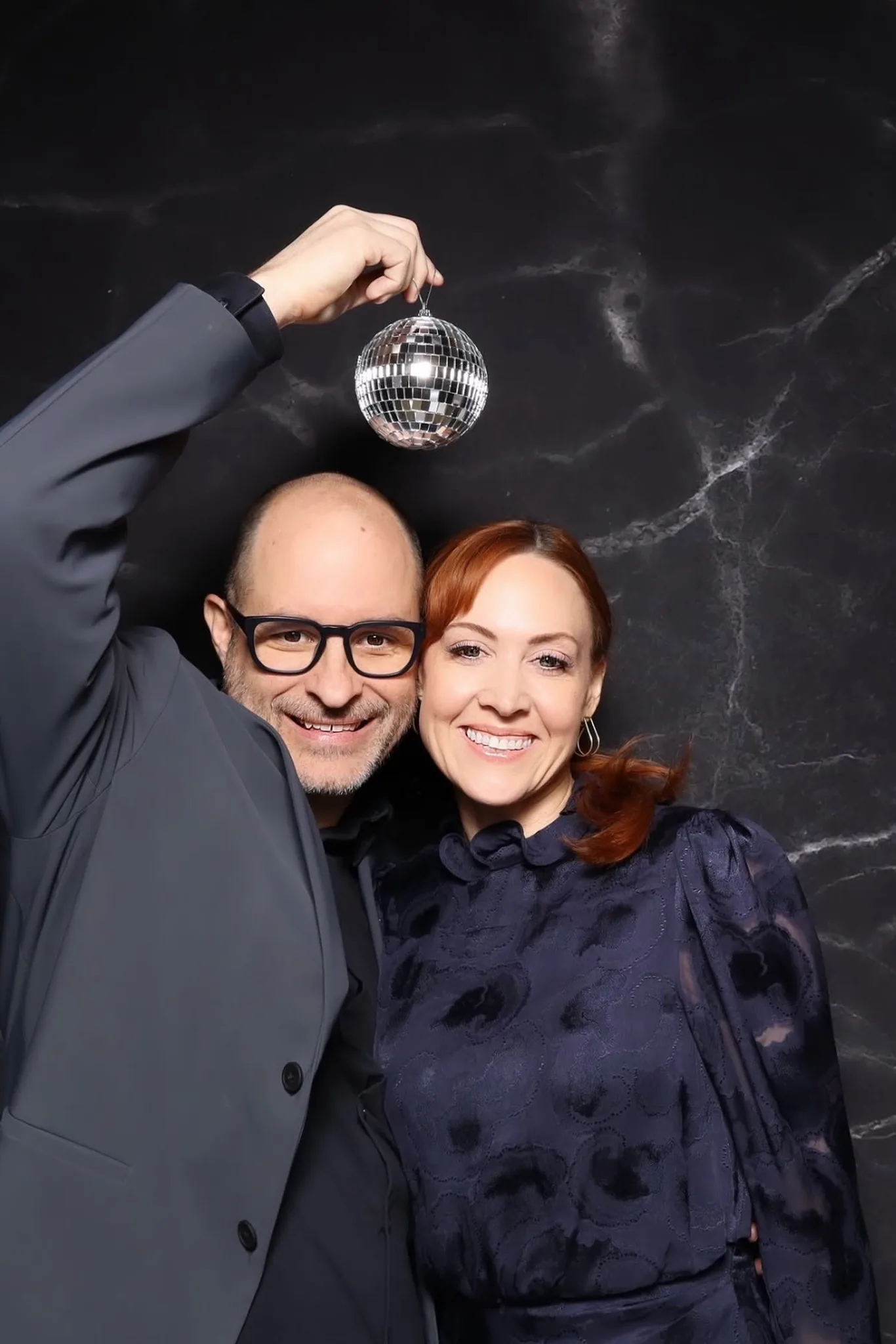 A couple posing with a disco ball in front of a black marble backdrop.